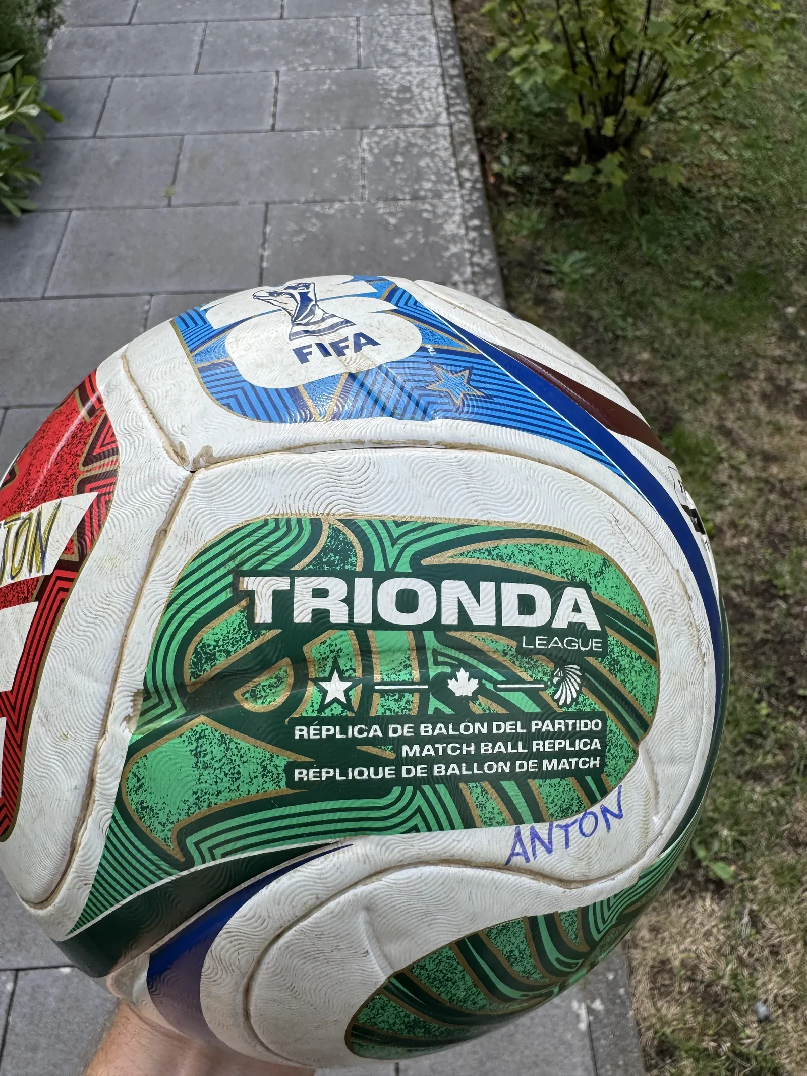 Weißer Fußball mit grünen, roten und blauen Feldern, in einer Hand über Pflastersteinen gehalten. Lesbar sind das FIFA-Zeichen, der Schriftzug TRIONDA LEAGUE und die Zeile MATCH BALL REPLICA. Auf ein weißes Feld ist mit blauem Filzstift ANTON geschrieben