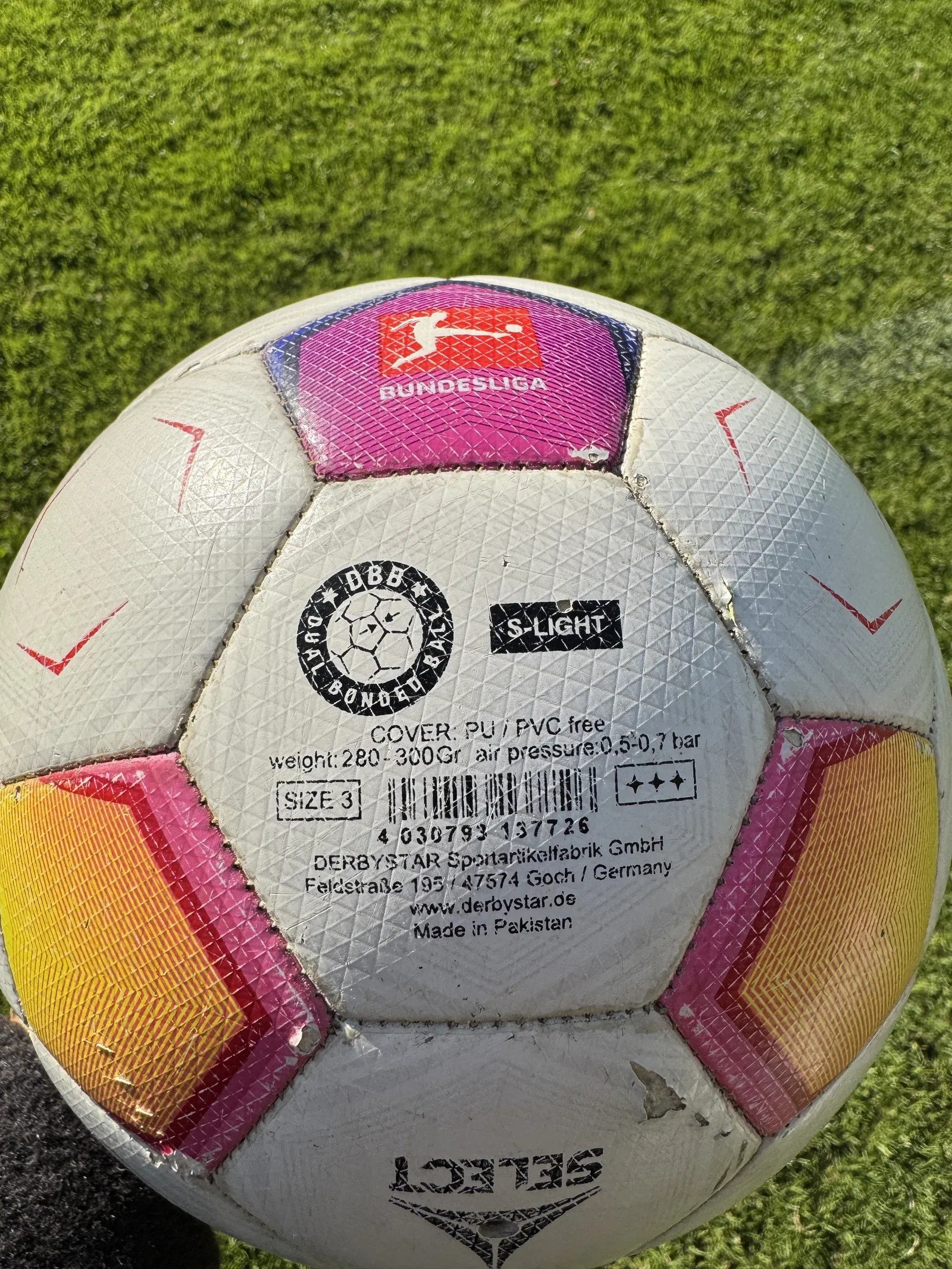 Nahaufnahme des Etiketts auf einem weiß-pink-orangen Fußball: Dual Bonded Ball, S-Light, Cover PU, weight 280 bis 300 Gramm, air pressure 0,5 bis 0,7 bar, SIZE 3
