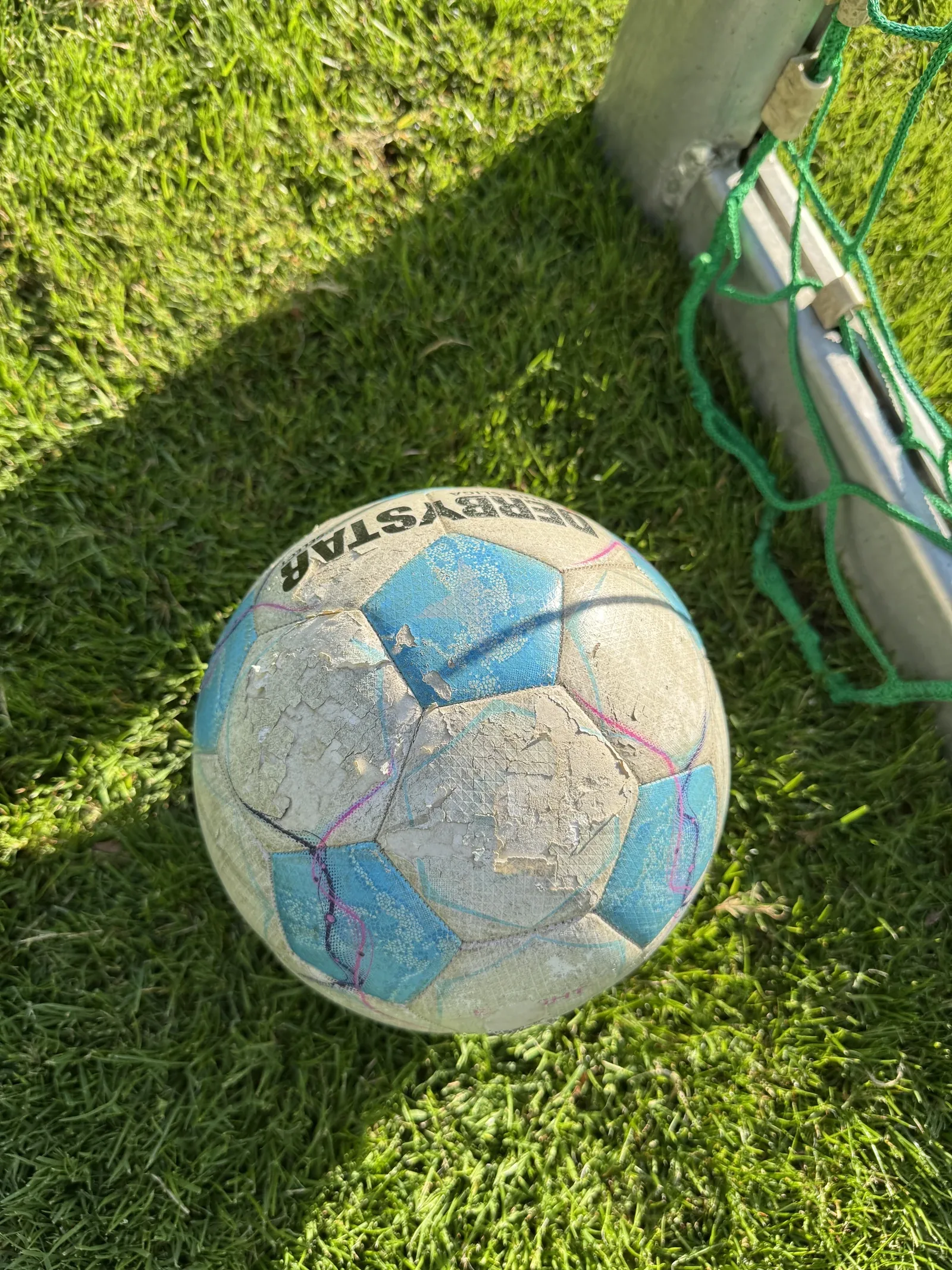 Blau-weißer Fußball in Größe 4 von oben im Gras neben dem Pfosten eines Jugendtors. Die weißen Felder der Hülle sind großflächig aufgeraut und ausgebrochen, der Aufdruck ist stellenweise verschwunden