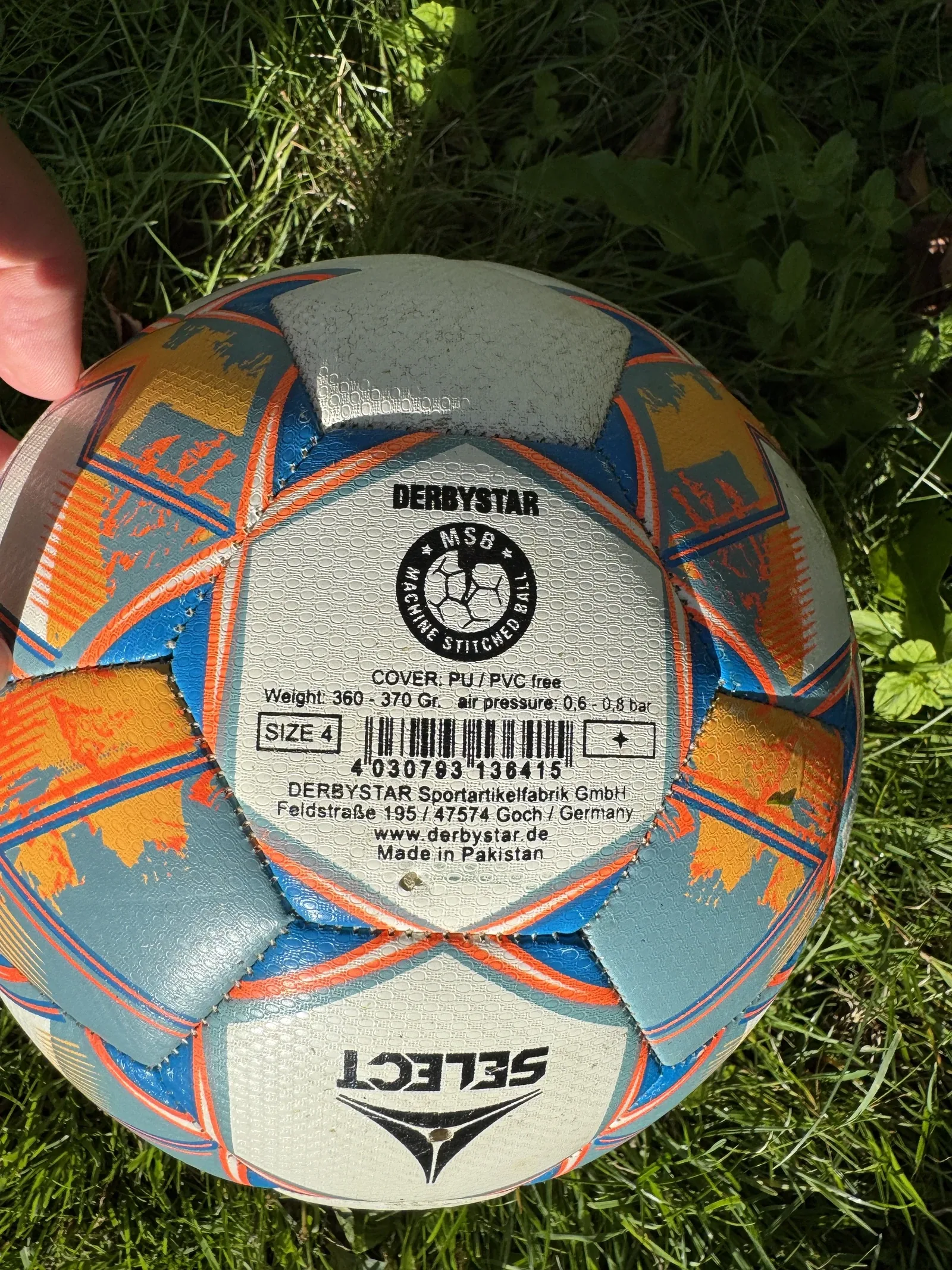 Nahaufnahme des Etiketts auf einem silber-blau-orangen Fußball, von einer Hand ins Bild gehalten. Lesbar sind DERBYSTAR, MSB Machine Stitched Ball, COVER PU PVC free, Weight 360 bis 370 Gr, air pressure 0,6 bis 0,8 bar und SIZE 4